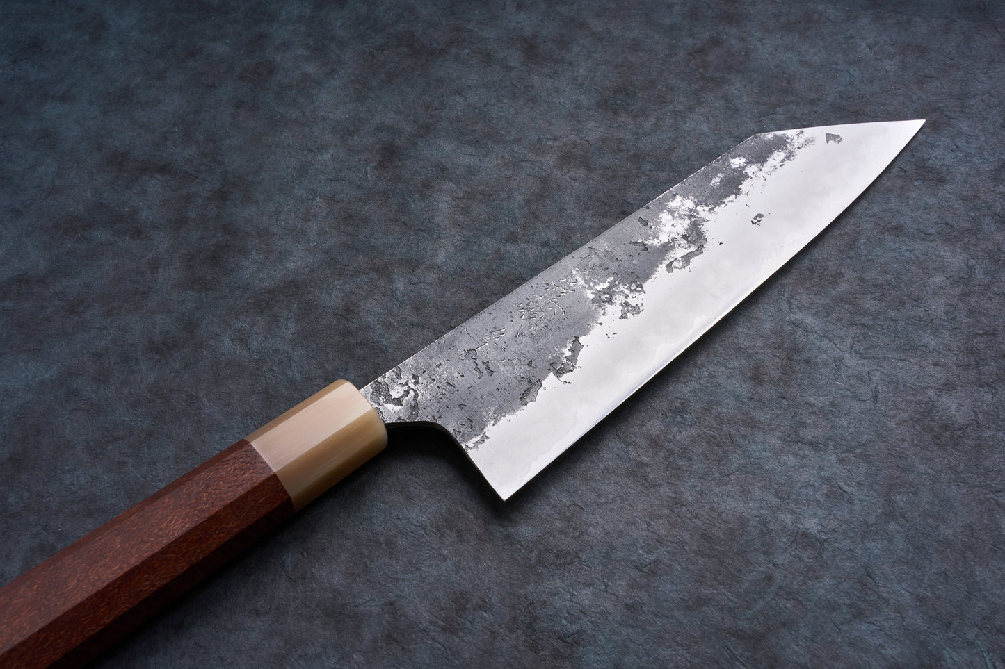 Mazaki Aogami Super Honsanmai Kurouchi Nashiji Kiritsuke Gyuto 210mm with a patina-finished steel blade and octagonal wooden handle on a stone surface.
