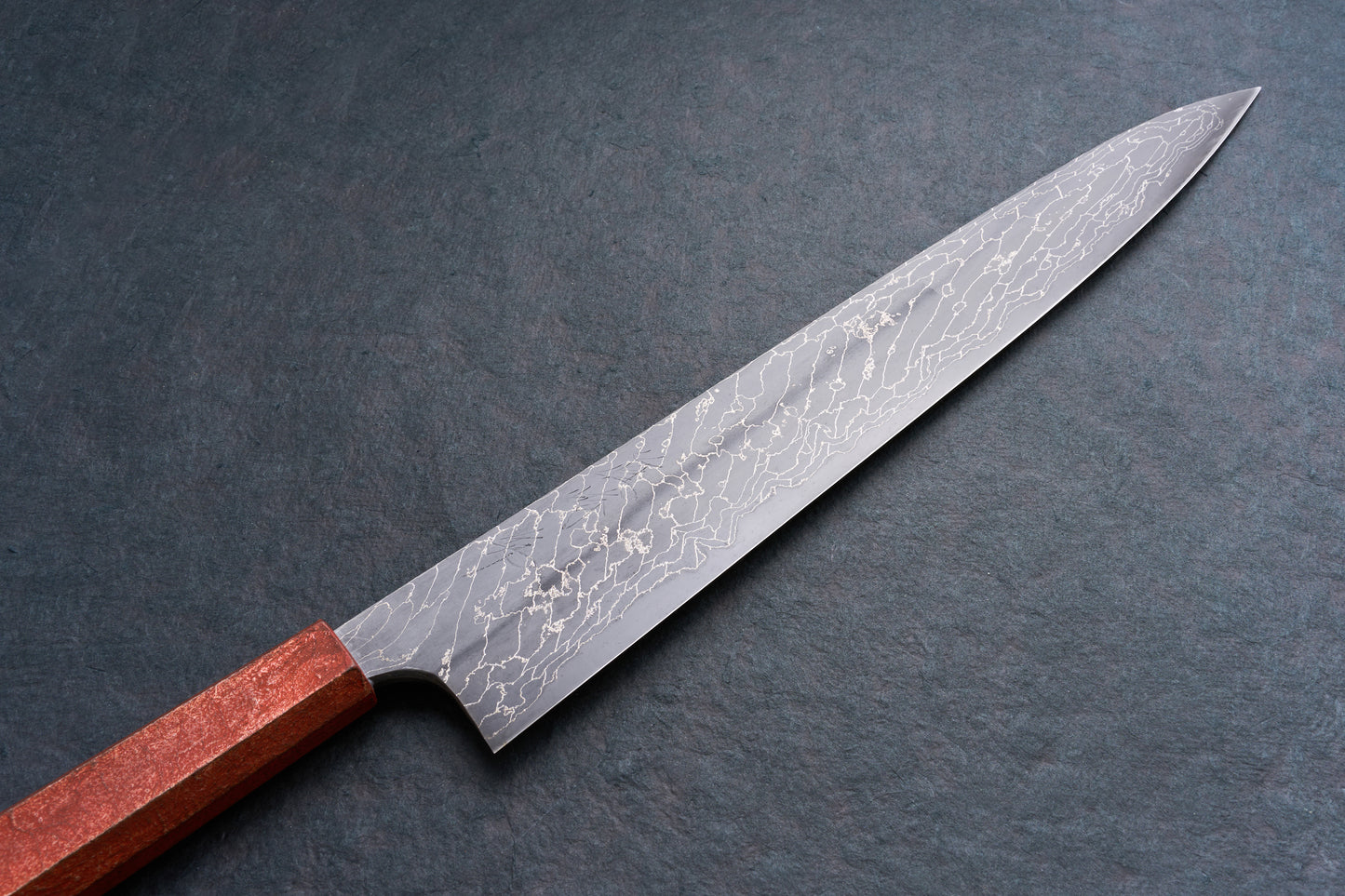 Mazaki Aoichi Beads Black Damascus Sujihiki 300mm, a Japanese slicing knife with a patterned blade and reddish wooden handle, on a dark surface.