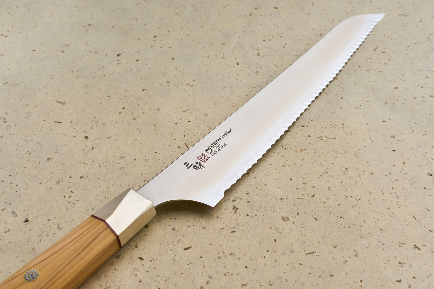 Mcusta Zanmai Beyond Bread Knife 230mm features a scalloped blade with Japanese markings, and an octagonal Hinoki wood handle, resting elegantly on a speckled stone surface, embodying Knifewear's dedication to exceptional Japanese craftsmanship.