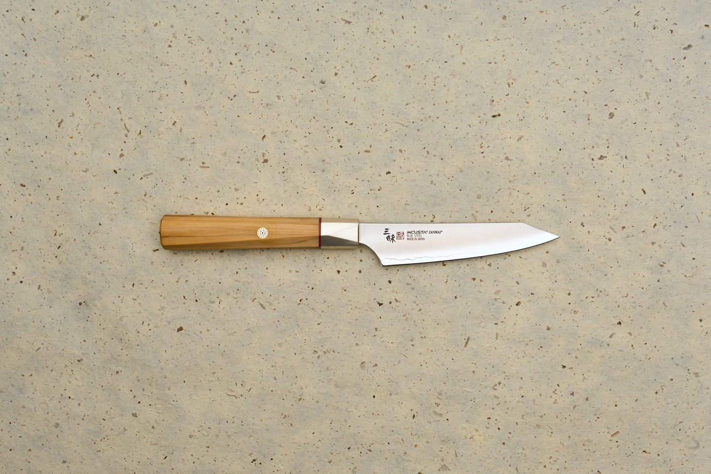 Mcusta Zanmai Beyond Kiritsuke Petty 110mm knife features a polished steel blade with MCUSTA ZANMAI branding and an octagonal Hinoki wood handle, laid on a speckled beige background.