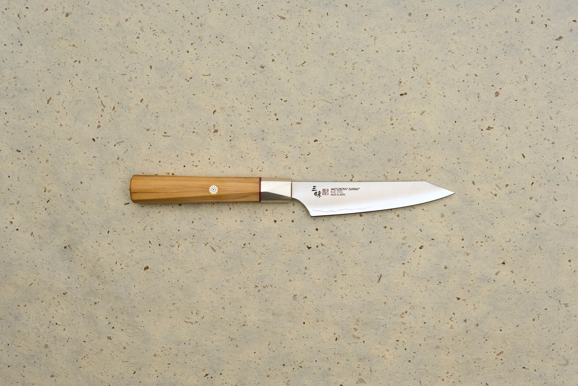 Mcusta Zanmai Beyond Kiritsuke Petty 110mm knife features a polished steel blade with MCUSTA ZANMAI branding and an octagonal Hinoki wood handle, laid on a speckled beige background.
