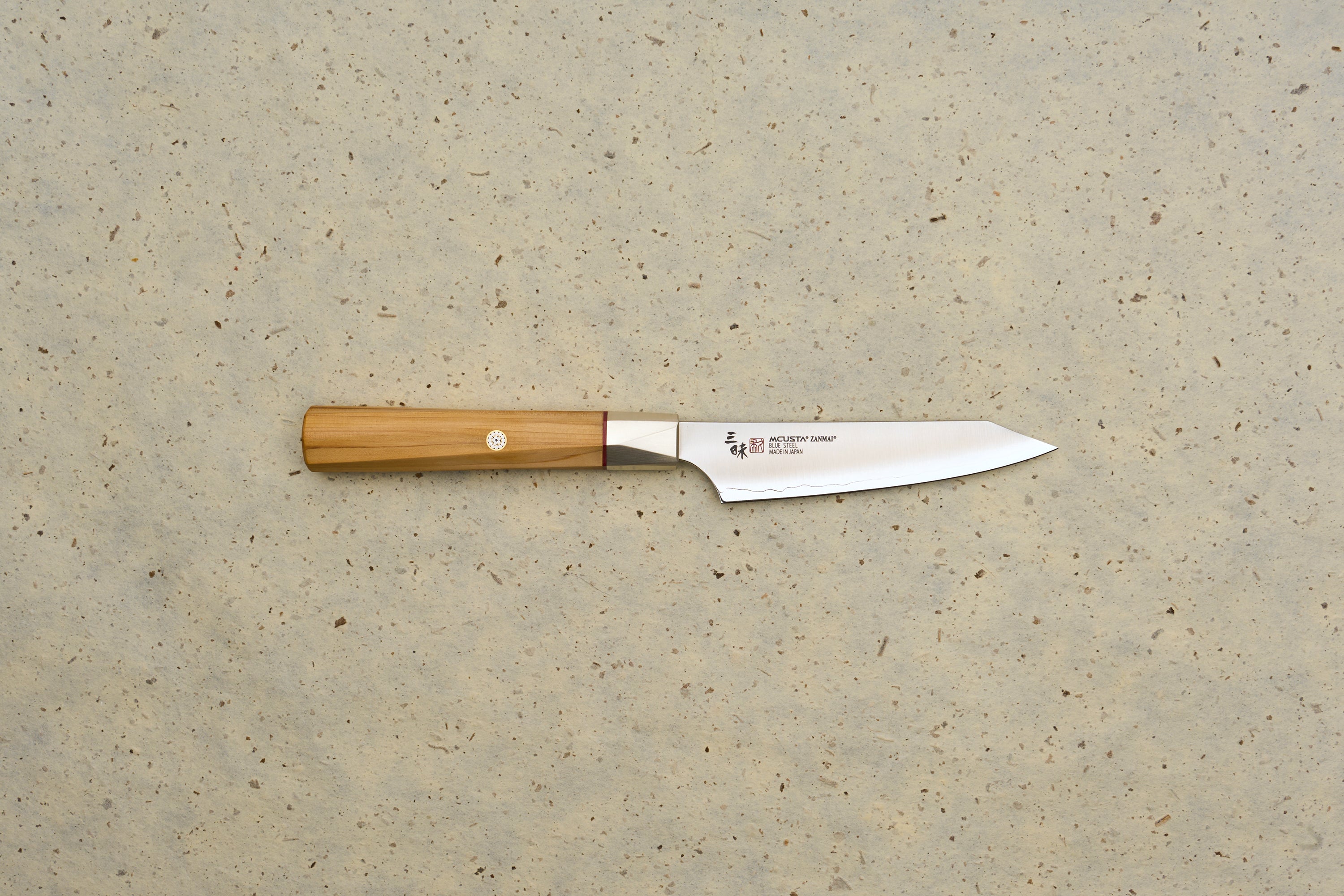 Mcusta Zanmai Beyond Kiritsuke Petty 110mm knife features a polished steel blade with MCUSTA ZANMAI branding and an octagonal Hinoki wood handle, laid on a speckled beige background.