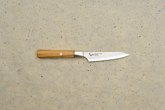 Mcusta Zanmai Beyond Kiritsuke Petty 110mm knife features a polished steel blade with MCUSTA ZANMAI branding and an octagonal Hinoki wood handle, laid on a speckled beige background.