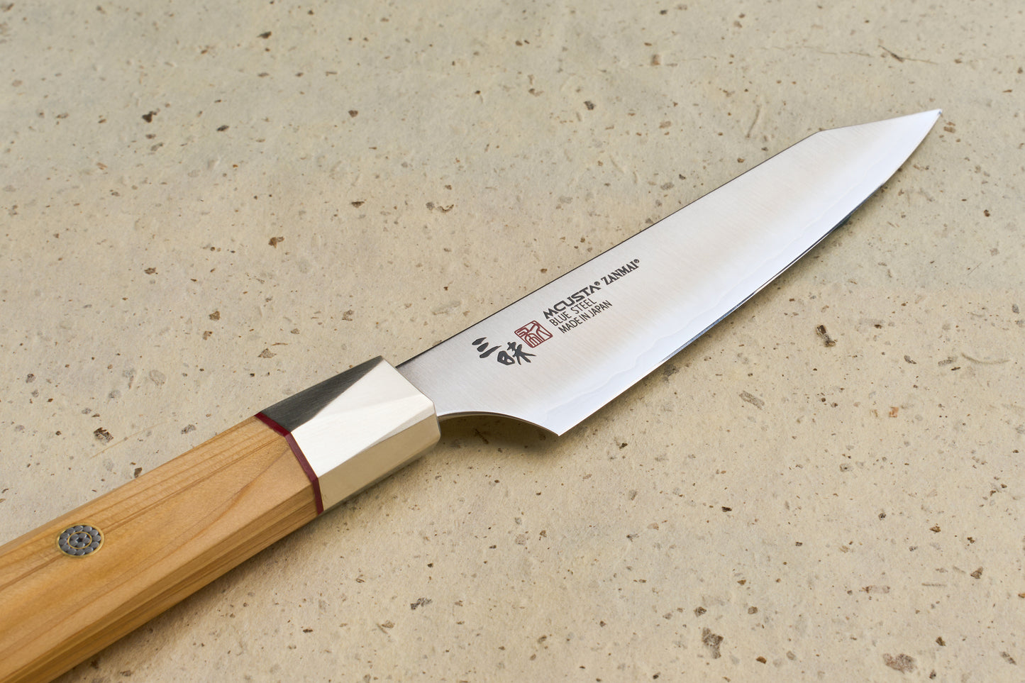 Mcusta Zanmai Beyond Kiritsuke Petty 110mm: A Japanese-style kitchen knife with a sleek blade, subtle hamon line, and octagonal Hinoki wood handle, resting on a speckled stone surface.