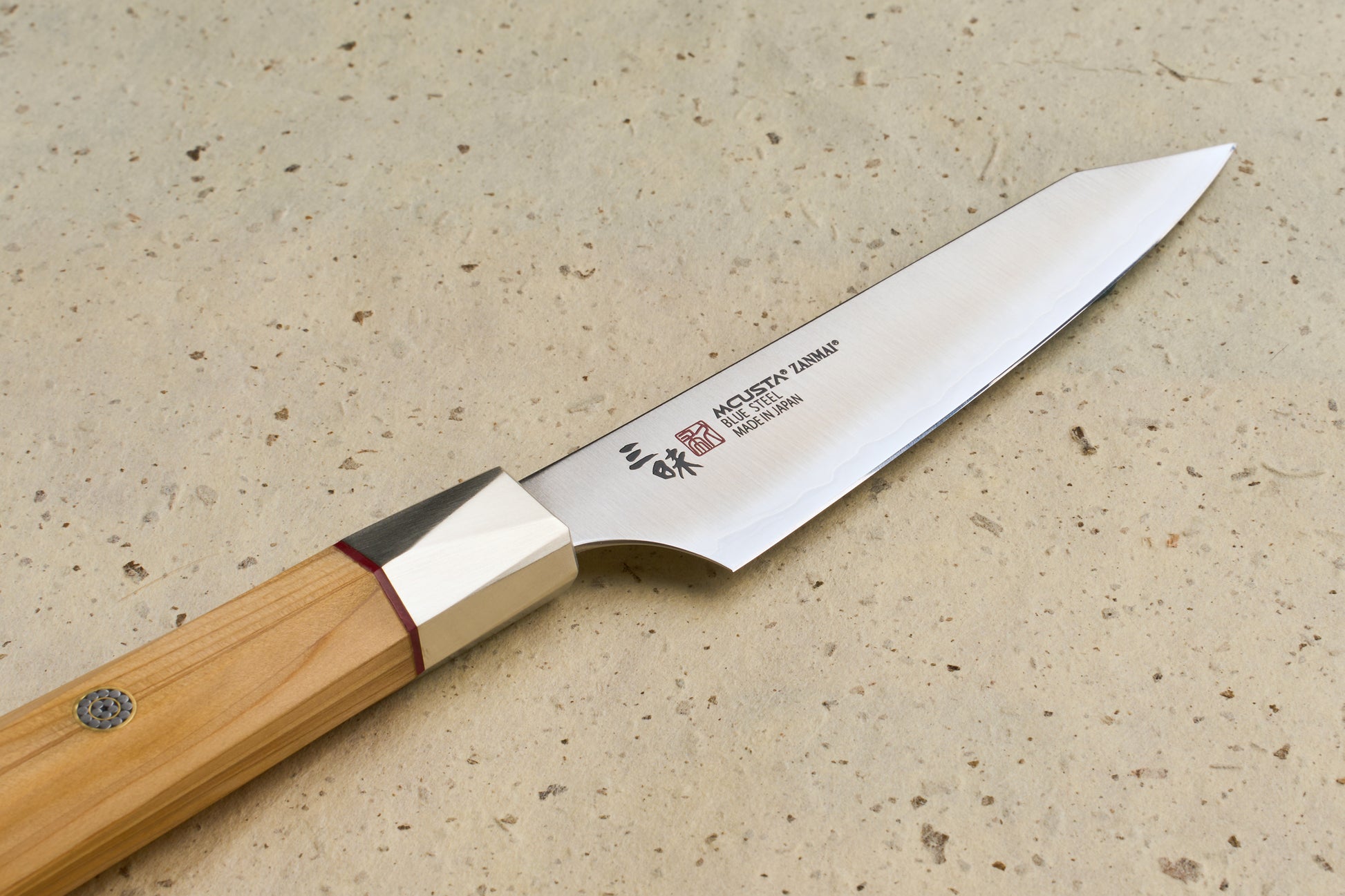 Mcusta Zanmai Beyond Kiritsuke Petty 110mm: A Japanese-style kitchen knife with a sleek blade, subtle hamon line, and octagonal Hinoki wood handle, resting on a speckled stone surface.