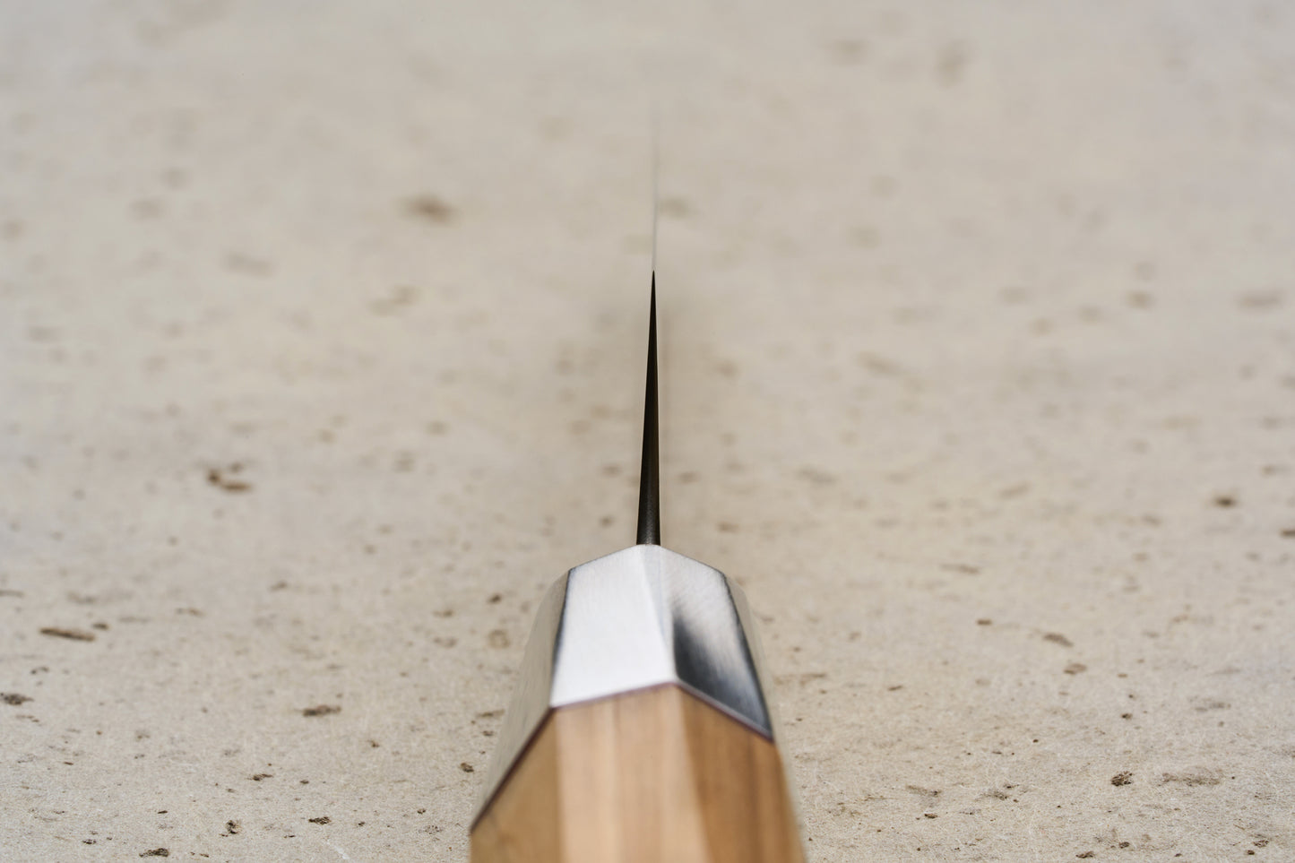 Close-up of the Mcusta Zanmai Beyond Kiritsuke Petty 110mm, featuring a thin metal blade from a geometric bolster and a faceted wooden handle, set against a speckled stone surface.