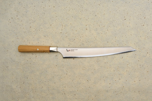 Mcusta Zanmai Beyond Kiritsuke Sujihiki 270mm knife on a stone surface, showcasing its high-carbon steel blade with hamon and octagonal wooden handle, featuring detailed maker's inscription and Japanese characters.
