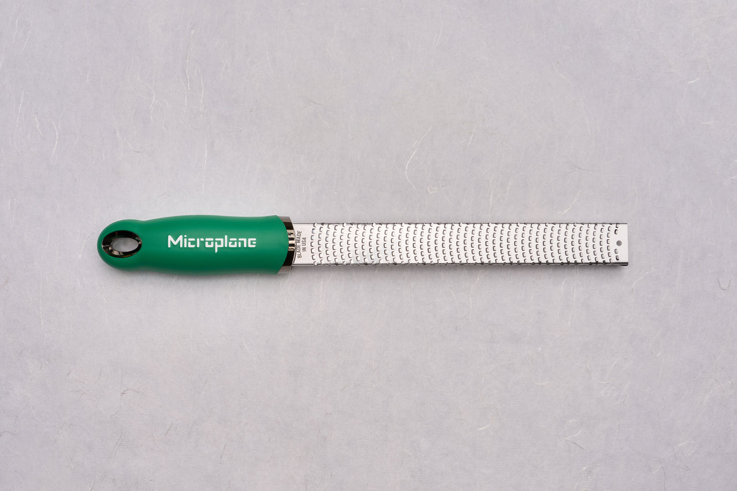 Microplane Premium Classic Zester with a green handle, featuring a narrow stainless-steel blade with fine, sharp teeth, ideal for zesting citrus and grating hard cheeses, embodying precision for culinary enthusiasts.