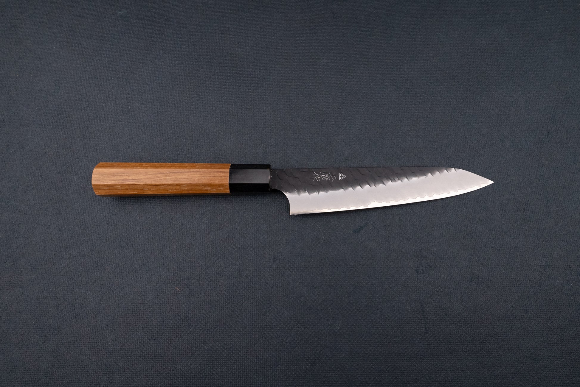 Nigara SG2 Kurouchi Tsuchime Petty 150mm knife features a long, gently curved blade with a hammered finish and an octagonal wooden handle, ideal for precise cutting tasks.