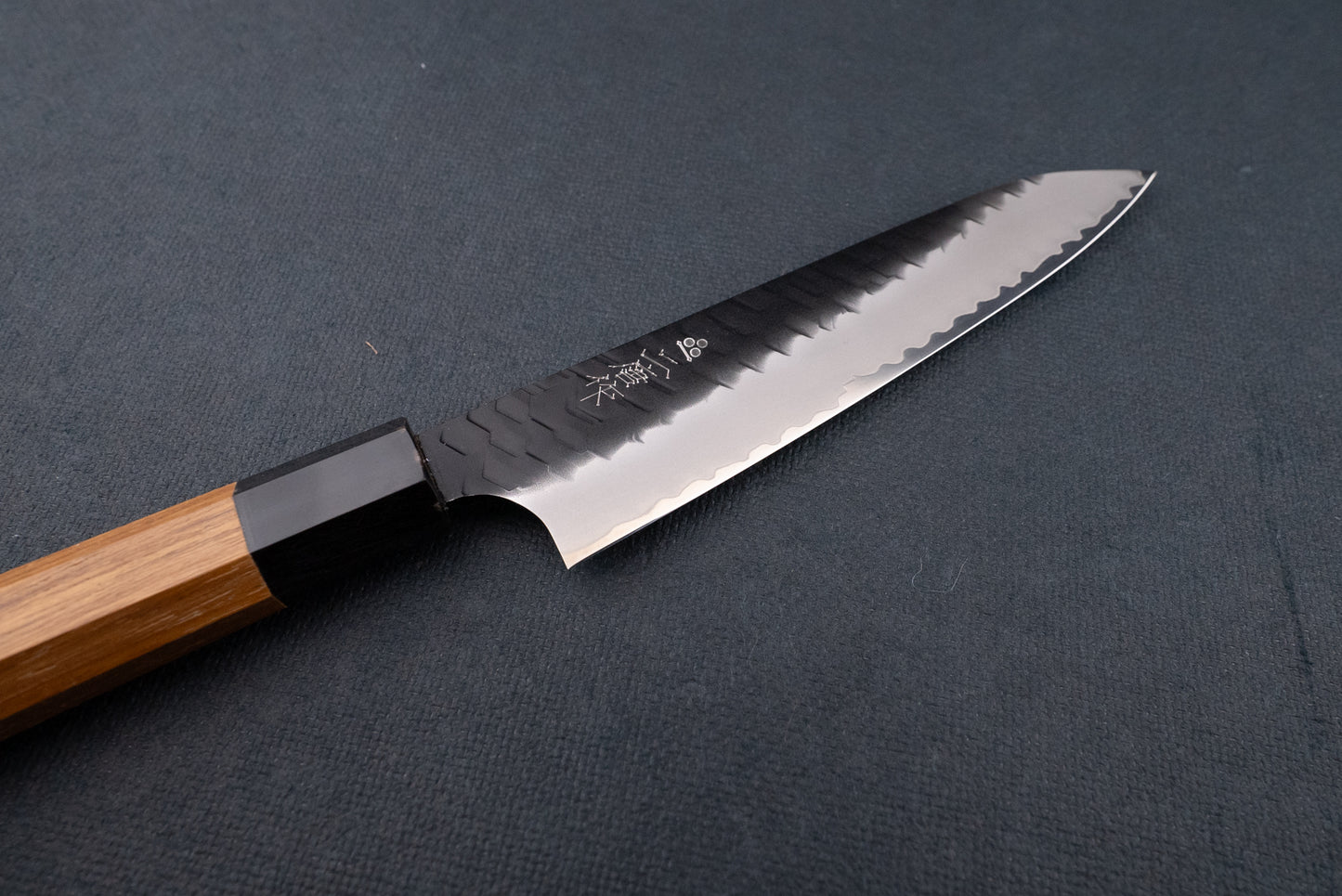 Nigara SG2 Kurouchi Tsuchime Petty 150mm knife with a patterned blade and engraved Japanese characters, featuring a light wood octagonal handle, displayed on a dark cloth background.