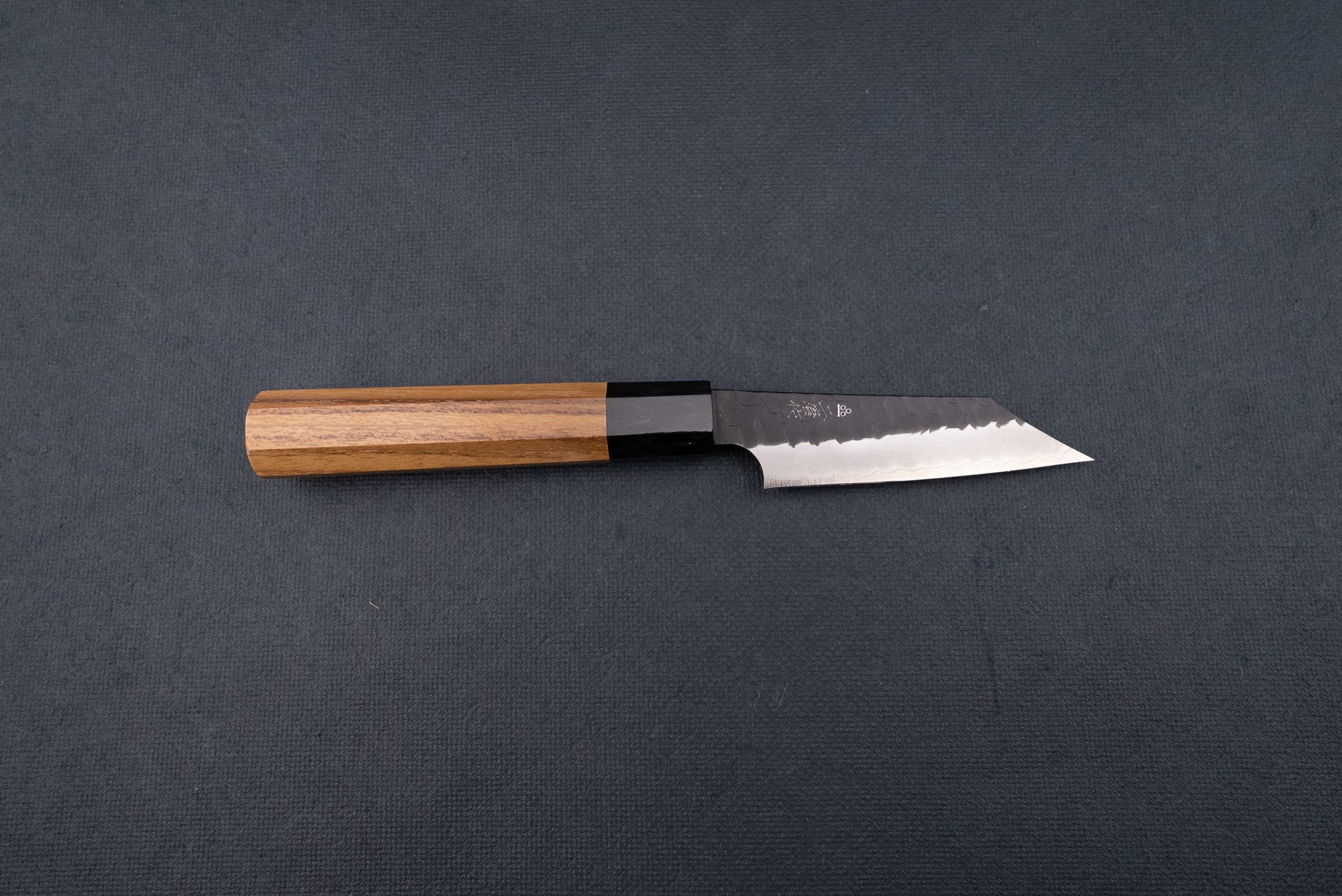 Nigara SG2 Kurouchi Tsuchime Kiritsuke Petty 95mm, a Japanese-style kitchen knife featuring a hammered single-bevel blade and octagonal wooden handle, displayed on a dark cloth backdrop.