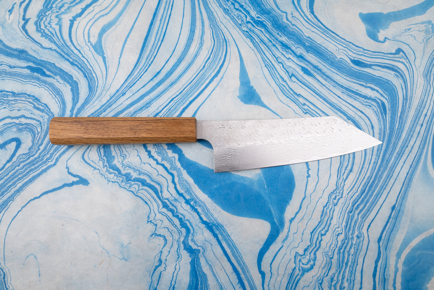Nigara SPG STRIX Tsuchime Damascus Bunka 180mm knife with a straight, octagonal wooden handle and a long, intricately patterned blade, displayed on a marbled surface, exemplifying Japanese craftsmanship.