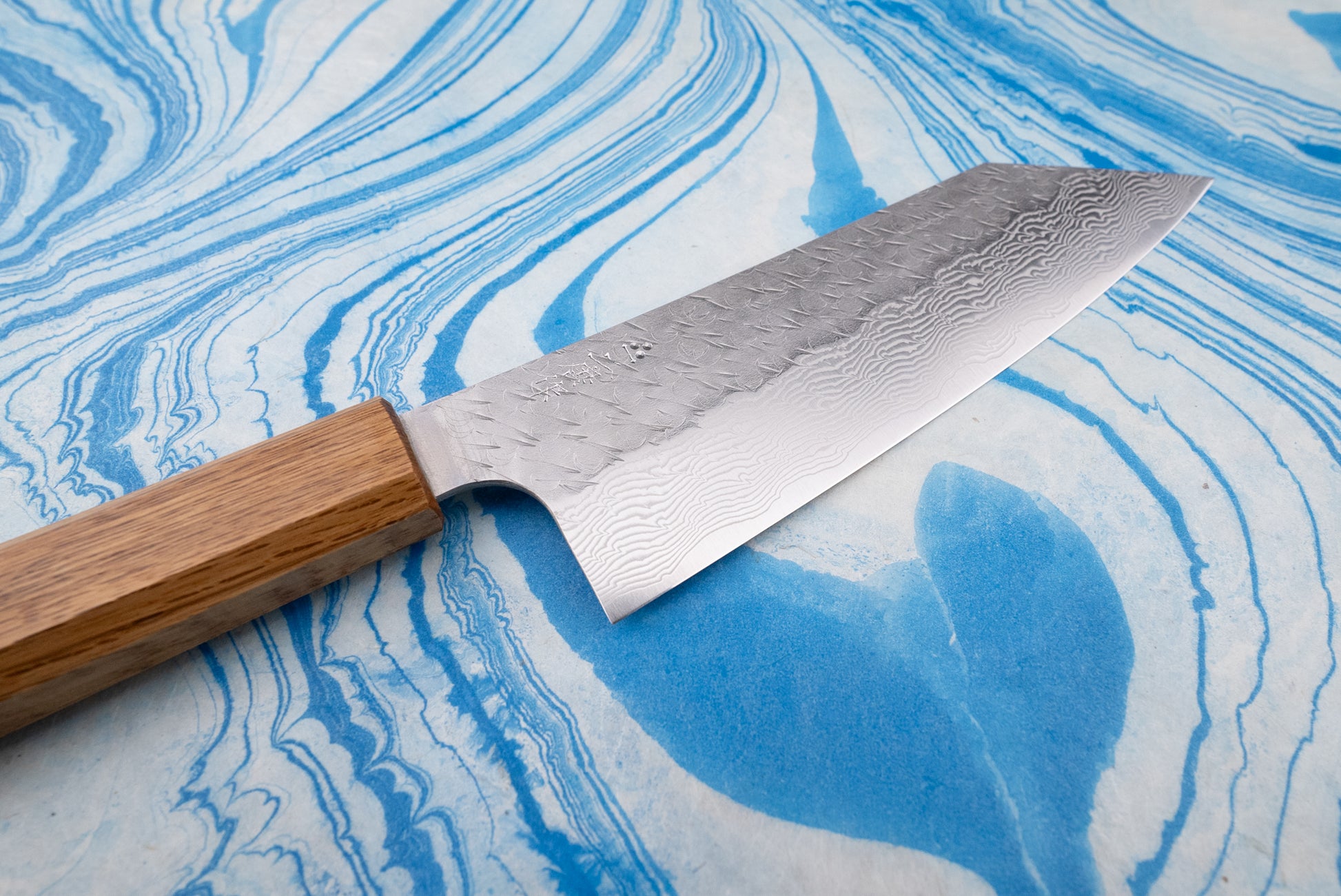 Nigara SPG STRIX Tsuchime Damascus Bunka 180mm knife on blue marbled surface, showcasing its Damascus-patterned blade with a hammered finish and an octagonal wooden handle, embodying Japanese craftsmanship.