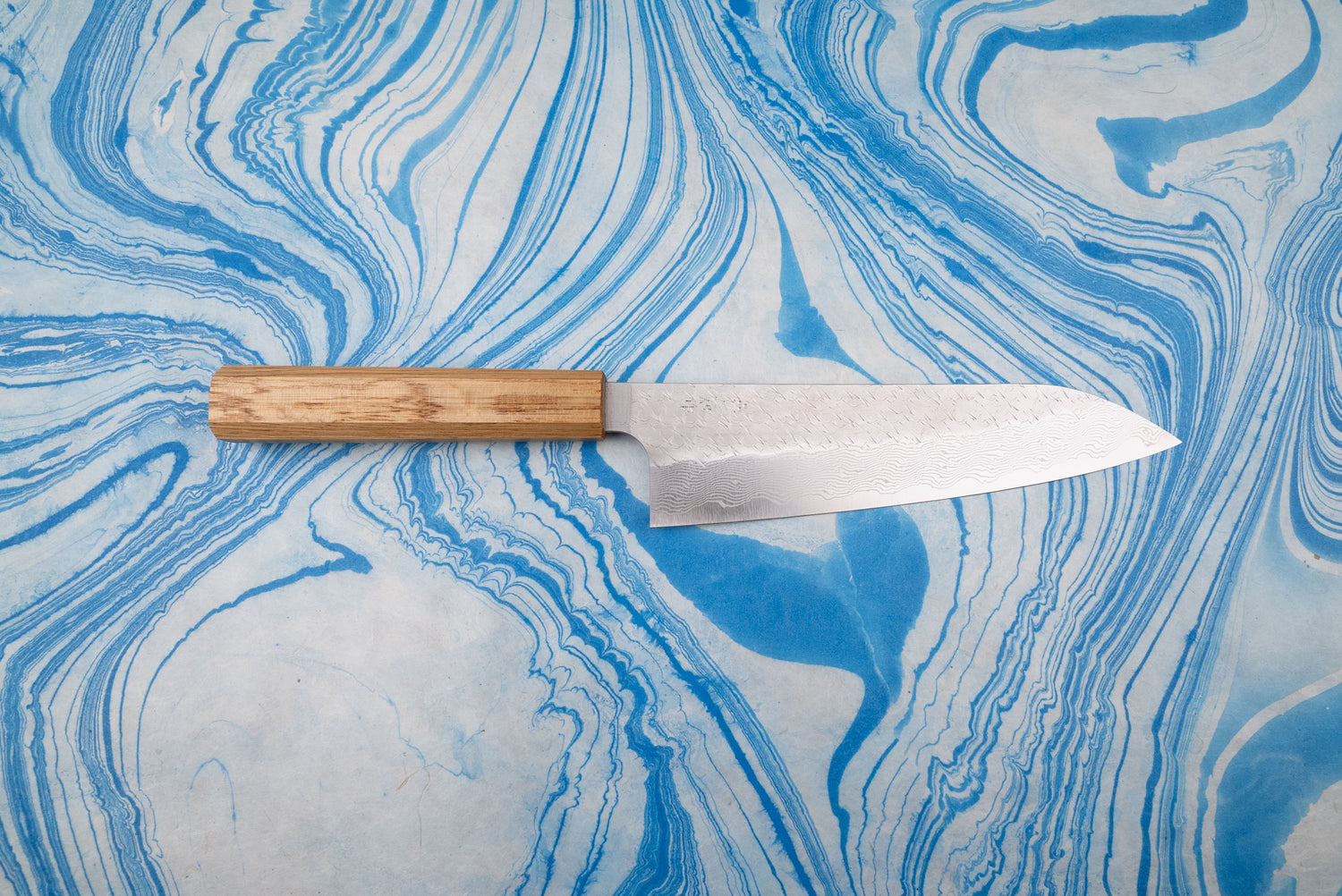Nigara SPG STRIX Tsuchime Damascus Gyuto 210mm knife with a wooden handle, showcasing a Damascus-style blade pattern, on a blue-and-white marbled surface, embodying Japanese craftsmanship and versatility.