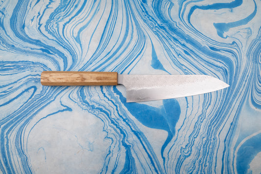 Nigara SPG STRIX Tsuchime Damascus Gyuto 210mm knife with a wooden handle, showcasing a Damascus-style blade pattern, on a blue-and-white marbled surface, embodying Japanese craftsmanship and versatility.