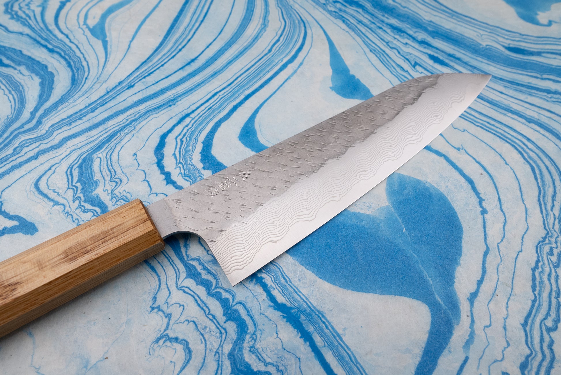 Nigara SPG STRIX Tsuchime Damascus Gyuto 210mm showcased with its exquisite Damascus steel blade and hand-hammered finish, featuring a light wooden octagonal handle, laid on a blue-and-white marbled surface.