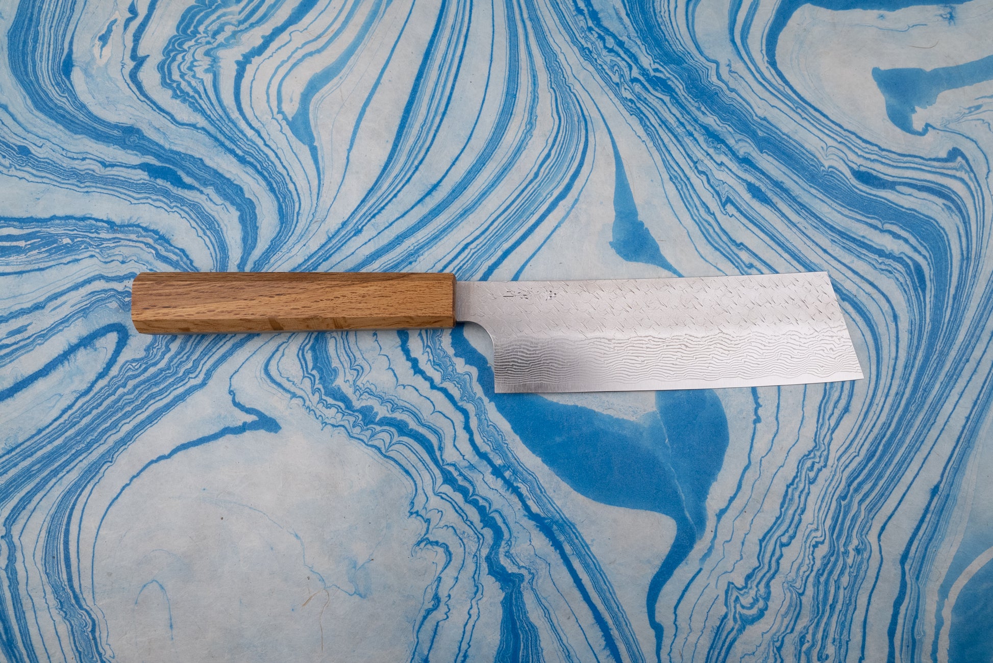 Nigara SPG STRIX Tsuchime Damascus Nakiri 180mm knife with a textured, layered blade pattern and natural wood handle, displayed on a blue-and-white marbled surface, exemplifying Japanese craftsmanship for vegetable chopping.