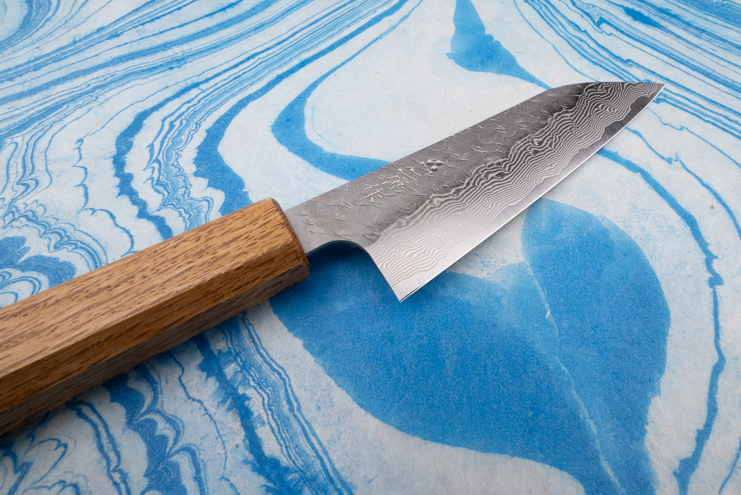 Nigara SPG STRIX Tsuchime Damascus Petty 120mm knife with a layered Damascus-steel pattern and rectangular wooden handle, set against a marbled blue-and-white background, showcasing its Japanese craftsmanship.