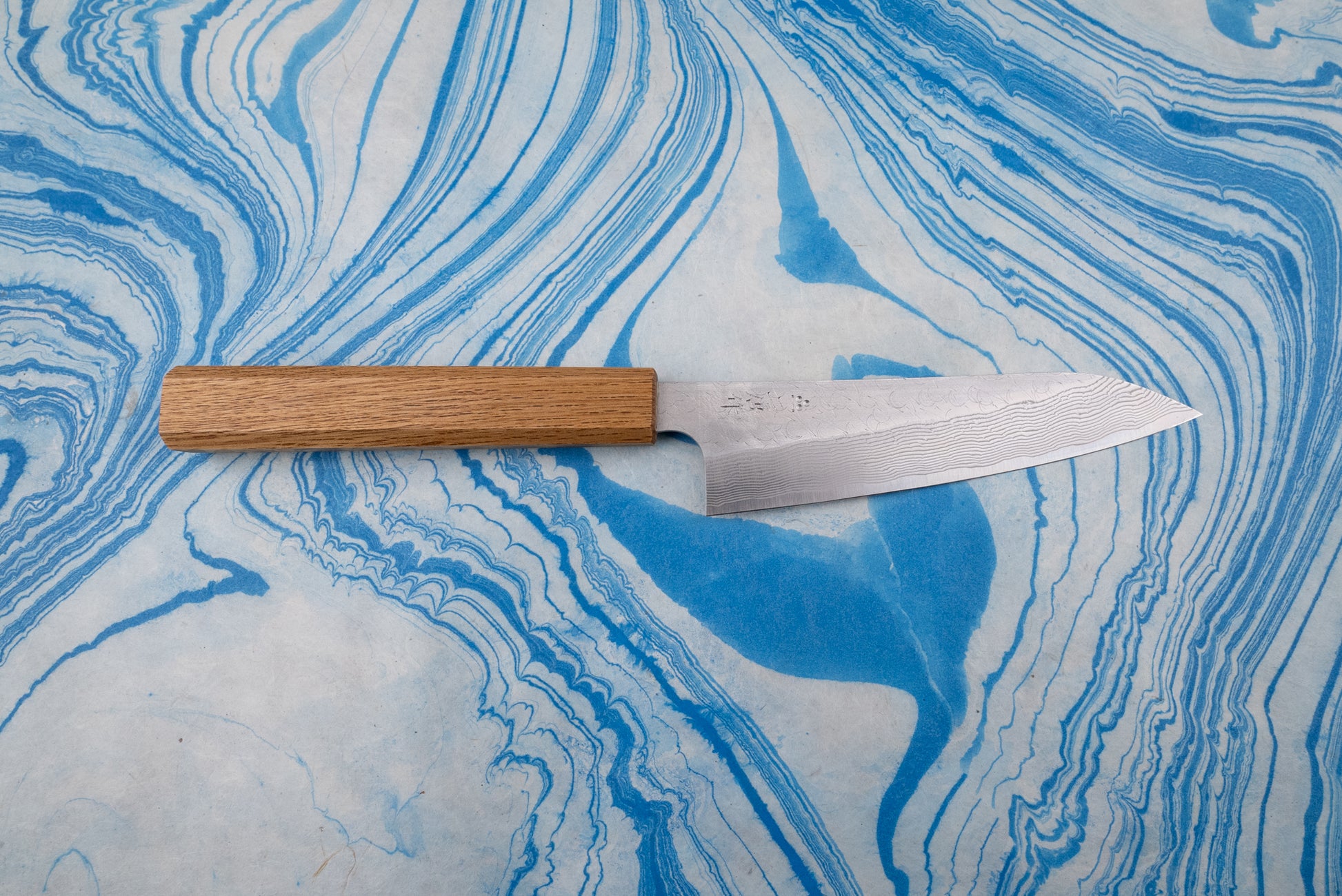 Nigara SPG STRIX Tsuchime Damascus Petty 150mm knife with a narrow blade featuring a subtle Damascus pattern and etched characters, paired with an octagonal wooden handle, lying on a marbled surface.