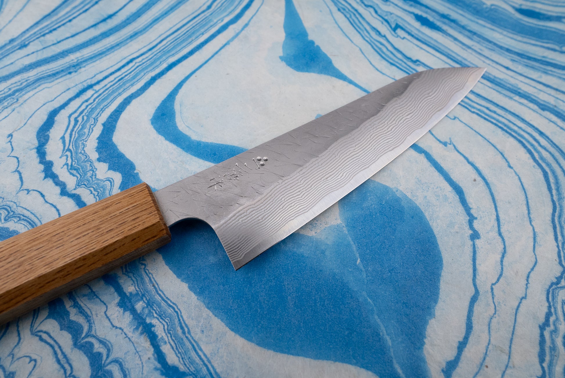 Nigara SPG STRIX Tsuchime Damascus Petty 150mm knife with a Japanese-style blade showcasing a Damascus-steel pattern and a wooden handle, resting on a blue marbled surface.