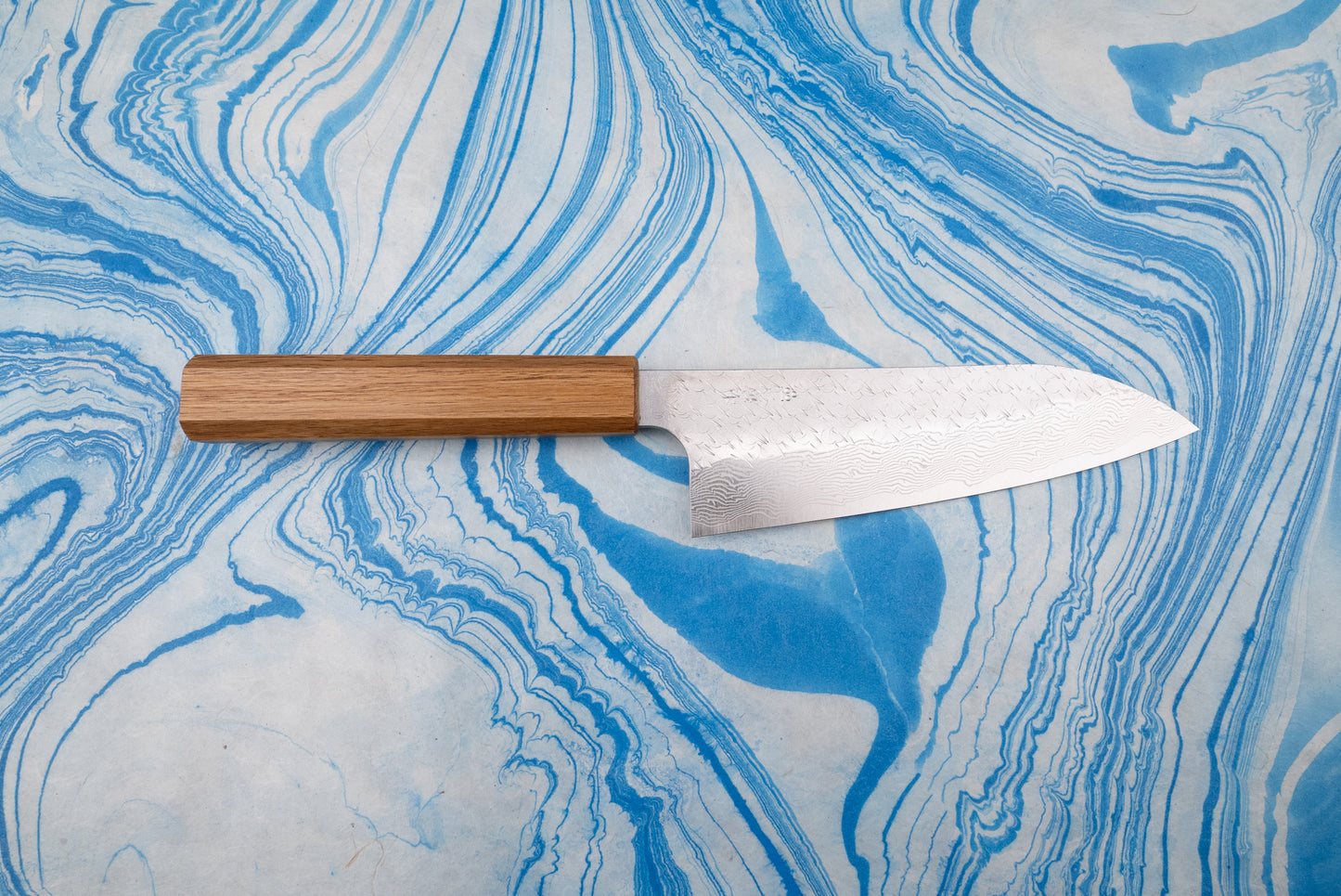 Nigara SPG STRIX Tsuchime Damascus Santoku 165mm knife on marbled surface, featuring a rectangular wooden handle and a wide, curved blade with intricate Damascus-style pattern, ideal for versatile kitchen use.