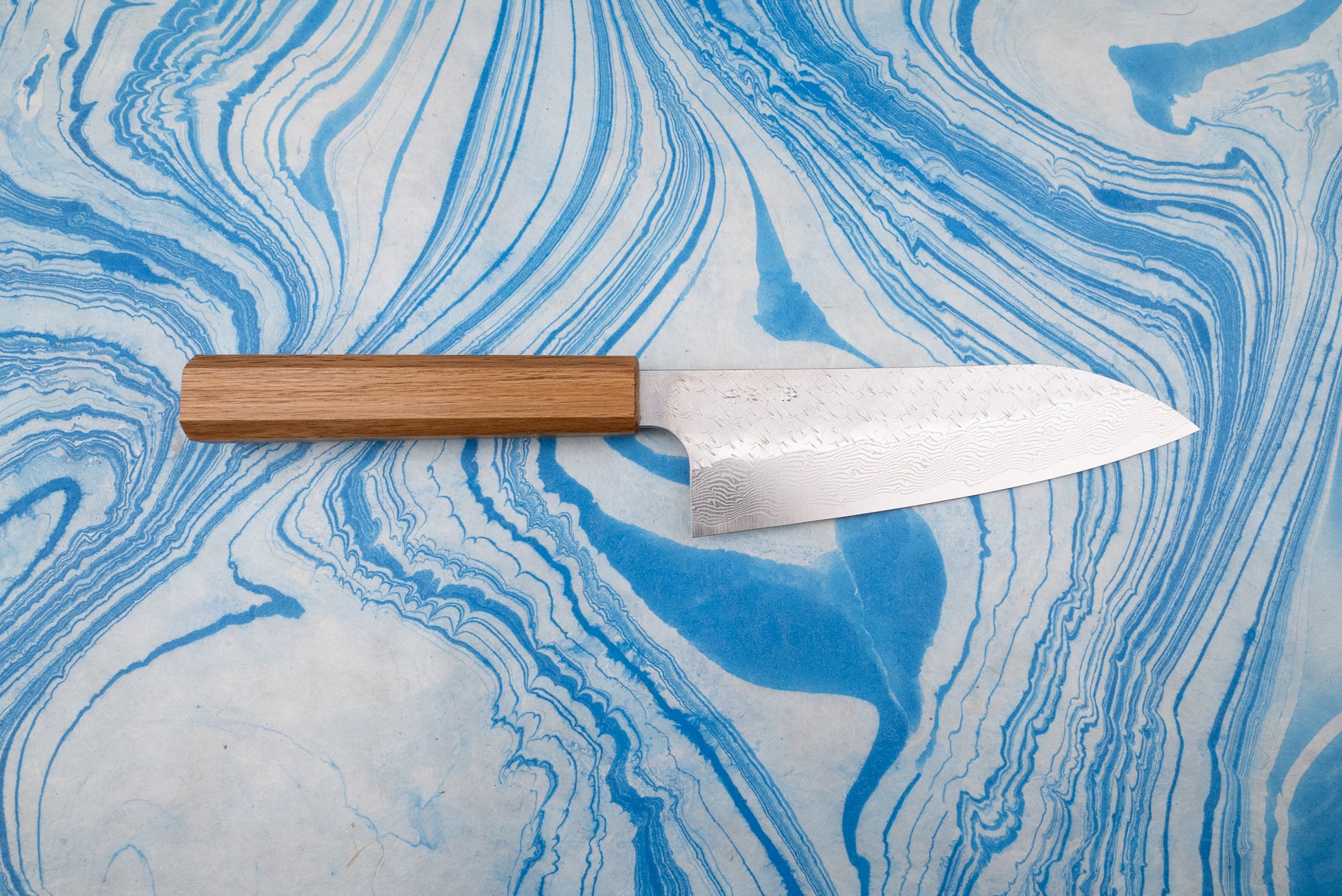 Nigara SPG STRIX Tsuchime Damascus Santoku 165mm knife on marbled surface, featuring a rectangular wooden handle and a wide, curved blade with intricate Damascus-style pattern, ideal for versatile kitchen use.