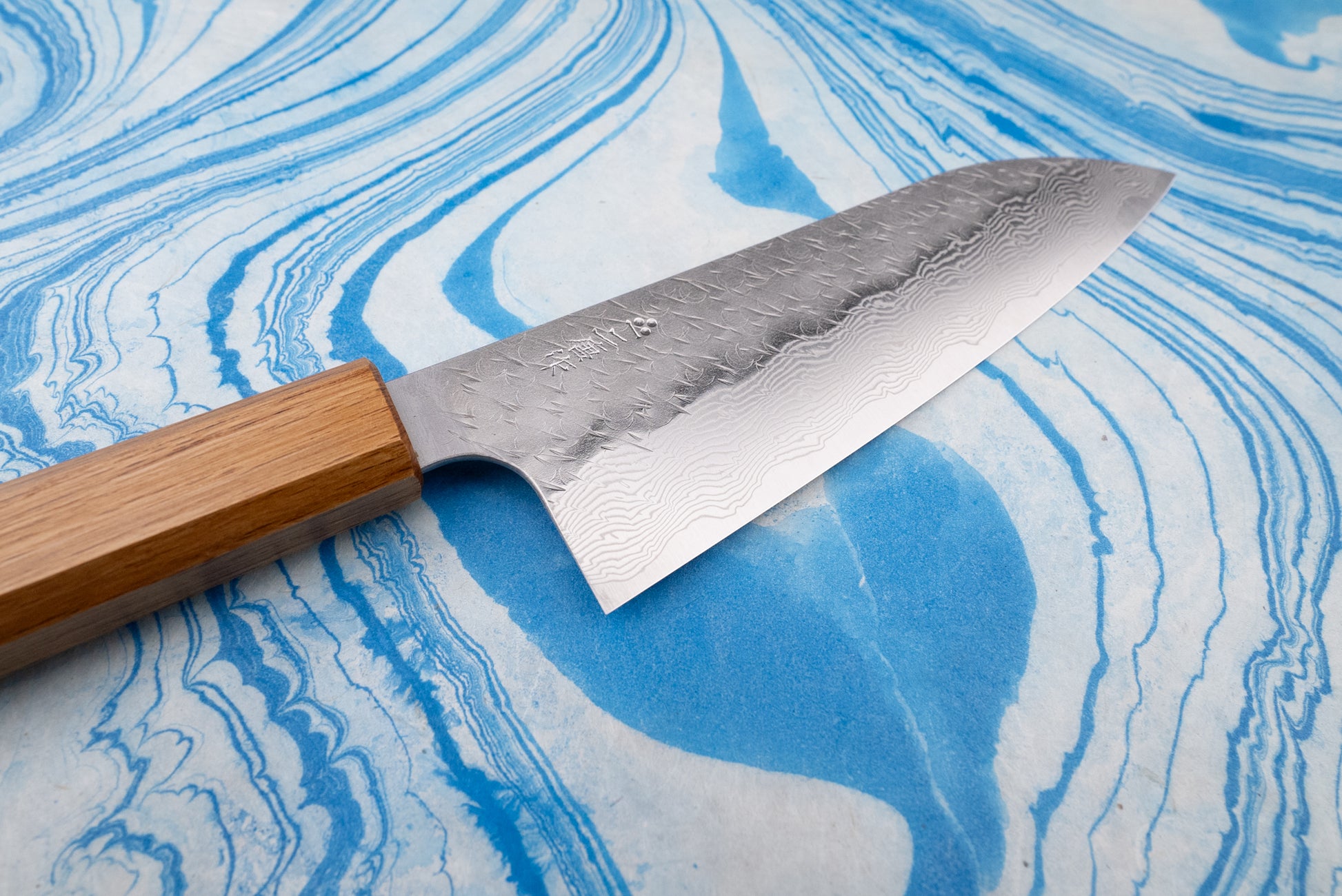 Nigara SPG STRIX Tsuchime Damascus Santoku 165mm, showcasing a Damascus-style patterned blade and an octagonal wooden handle, lies on a marbled surface, embodying Nigara Hamono's rich knifemaking tradition.