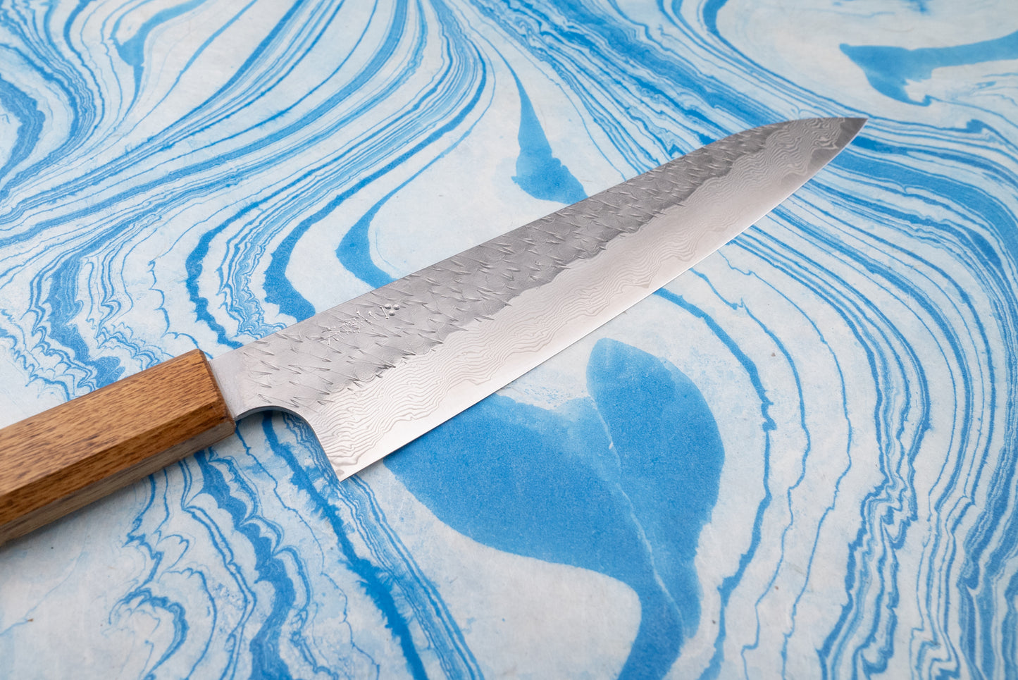 Nigara SPG STRIX Tsuchime Damascus Sujihiki 240mm knife with a hammered, Damascus-style blade and wooden handle, displayed on a blue marbled surface, embodies Japanese craftsmanship from Aomori's esteemed knifemaker.