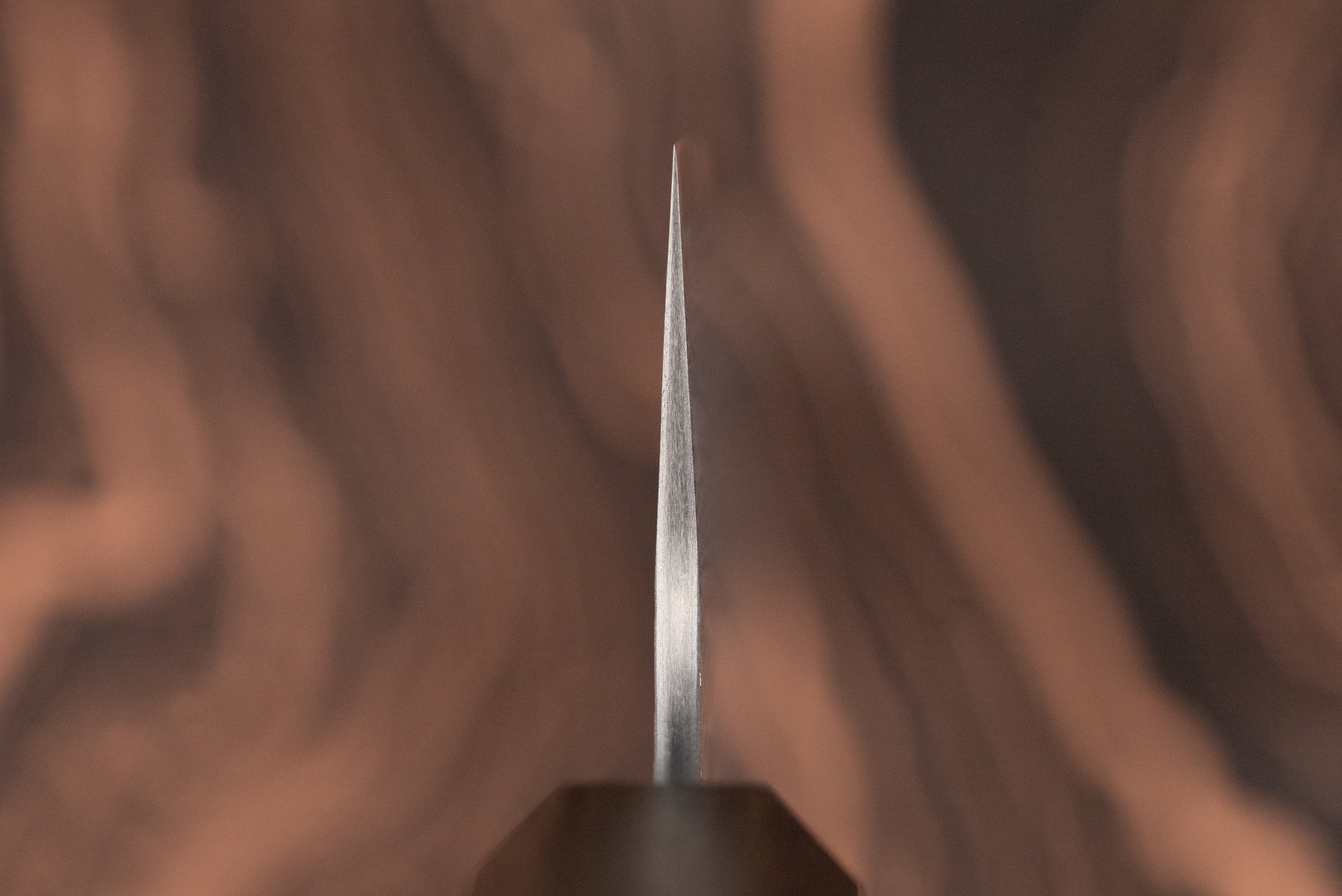 Macro shot of the Nigara VG XEOS Tsuchime Damascus Gyuto 225mm blade, showcasing its thin spine and sharp edge against a softly blurred wood-grain background, epitomizing Japanese craftsmanship.