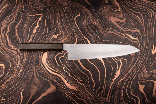 Nigara VG XEOS Tsuchime Damascus Gyuto 255mm with a damascus-patterned blade and dark wooden octagonal handle on a marbled, copper-accented surface, exemplifying Japanese craftsmanship from Aomori's historic knifemaker.