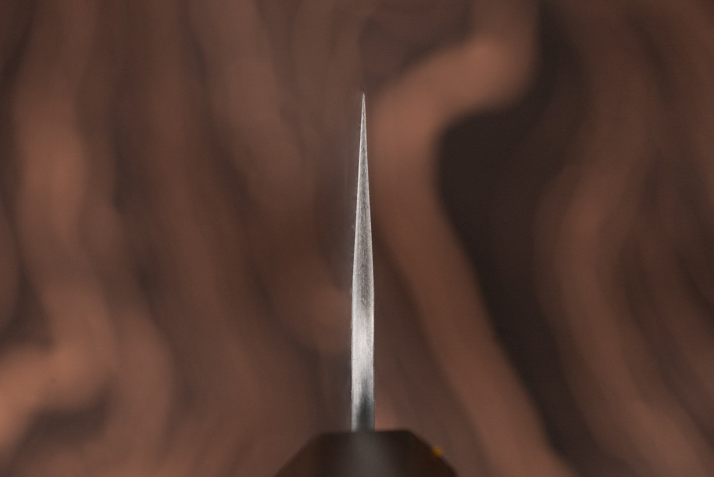 Close-up of the sharp tip of a Nigara VG XEOS Tsuchime Damascus Gyuto 255mm knife, showcasing its finely ground edge against a blurred wood-grain background, reflecting exquisite Japanese craftsmanship.