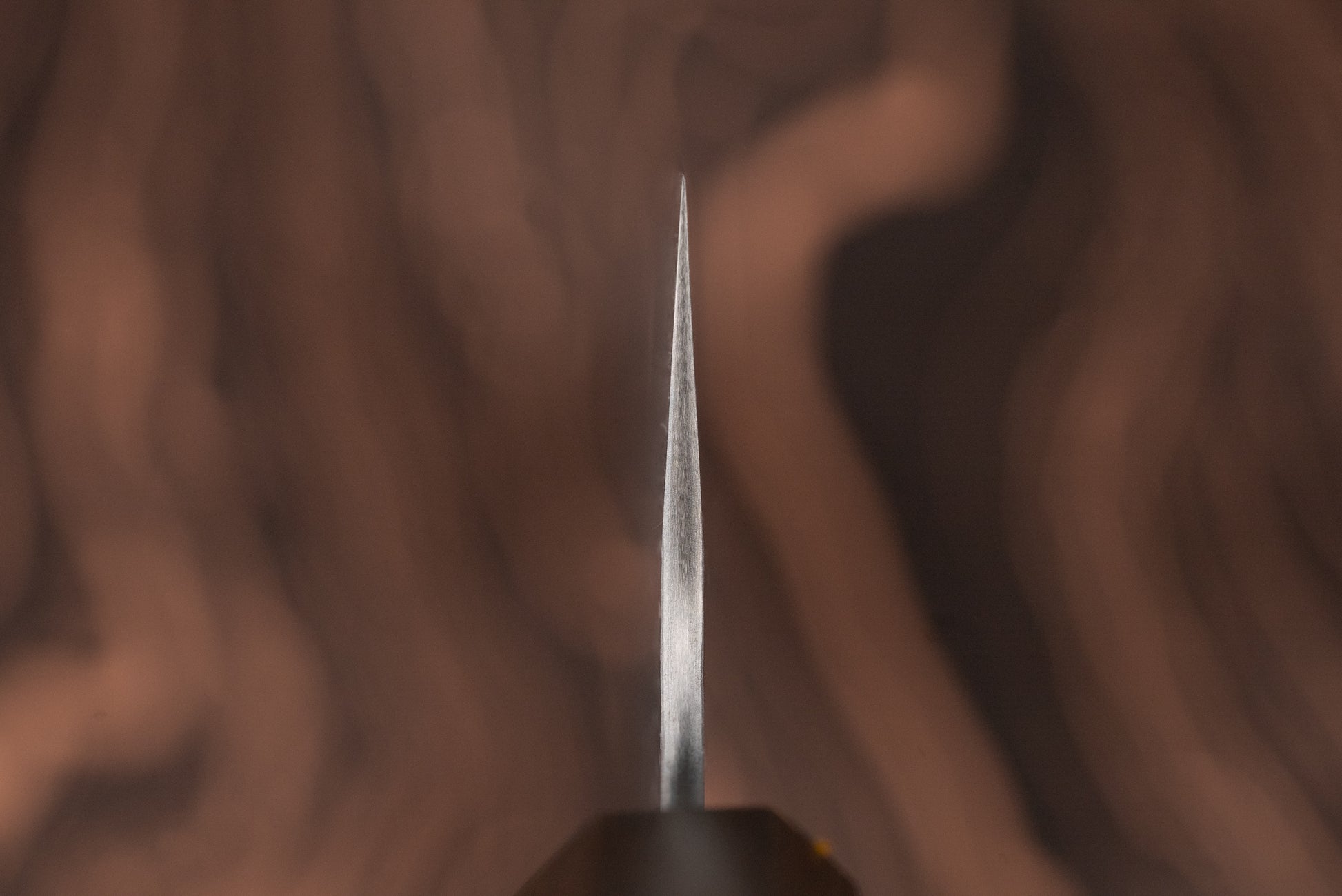 Close-up of the sharp tip of a Nigara VG XEOS Tsuchime Damascus Gyuto 255mm knife, showcasing its finely ground edge against a blurred wood-grain background, reflecting exquisite Japanese craftsmanship.
