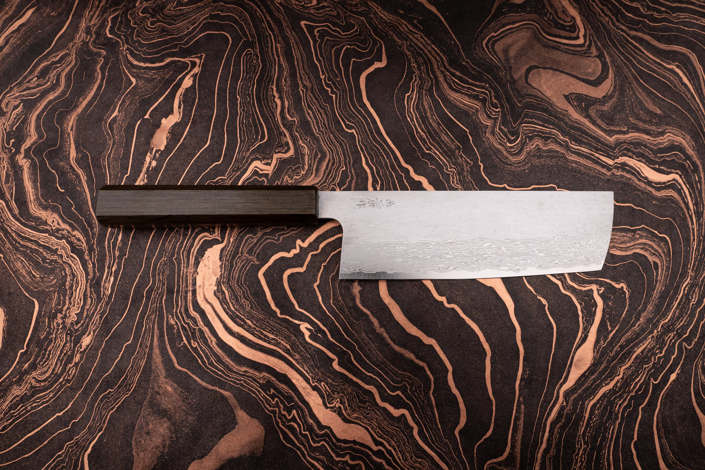 Nigara VG XEOS Tsuchime Damascus Nakiri 180mm, featuring a straight-edged Damascus-pattern blade and octagonal dark wooden handle, displayed on a marbled coppery surface. Ideal for efficient vegetable chopping.