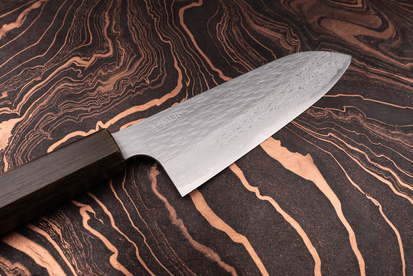 Nigara VG XEOS Tsuchime Damascus Santoku 185mm knife with a patterned steel blade and dark wooden handle, showcased on a marbled background, highlighting its artisanal craftsmanship from Aomori, Japan.