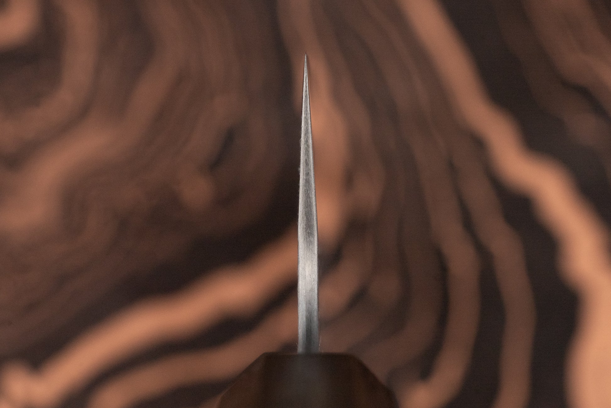 Macro shot of the Nigara VG XEOS Tsuchime Damascus Santoku 185mm blade, showcasing its fine-tapered tip against a blurred wood-grain pattern, highlighting its precision craftsmanship from Aomori's esteemed knifemaker.