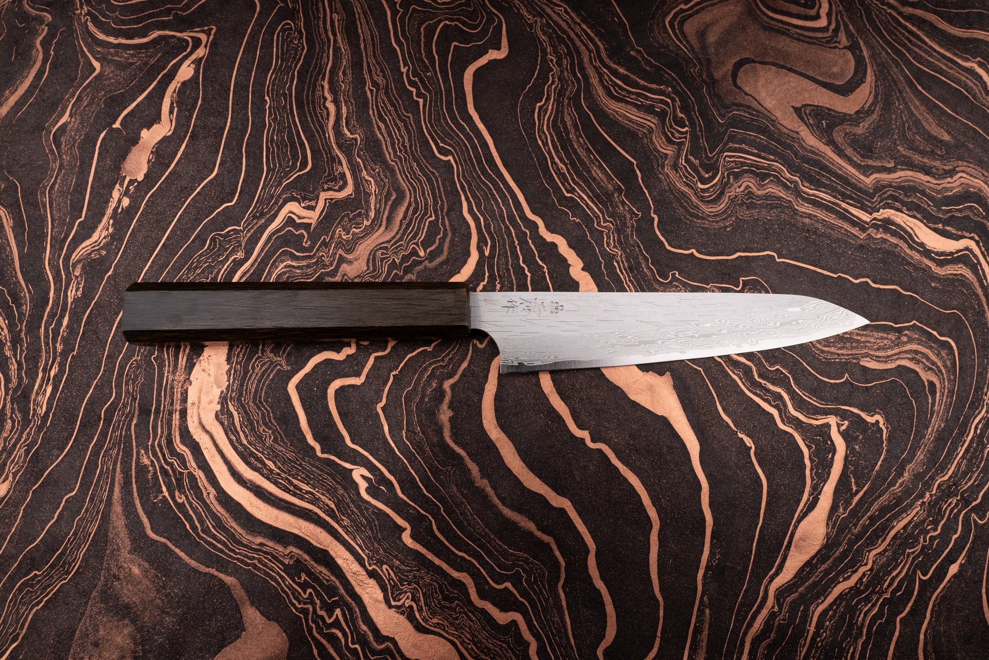 Nigara VG XEOS Tsuchime Damascus Petty 160mm knife with a slender damascus-patterned blade and dark wooden handle, positioned on a dark, marbled surface, showcasing its elegant, handcrafted design.