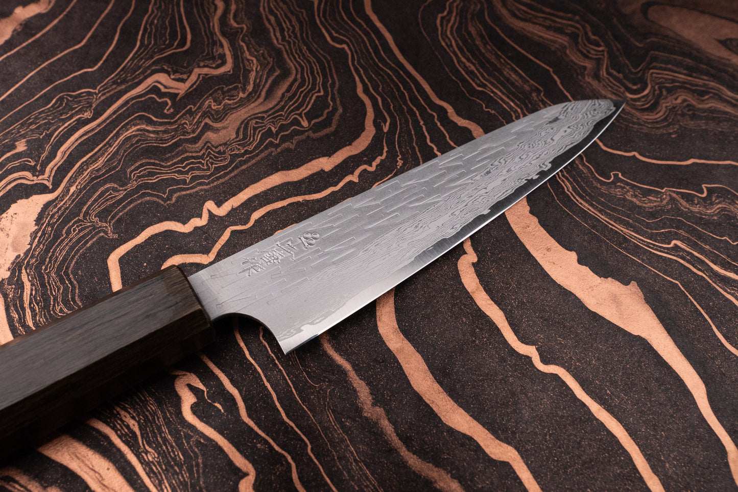 Nigara VG XEOS Tsuchime Damascus Petty 160mm knife with a Damascus-style, hammered blade and dark octagonal wooden handle, displayed on a black-and-copper marbled surface, ideal for precise kitchen tasks.