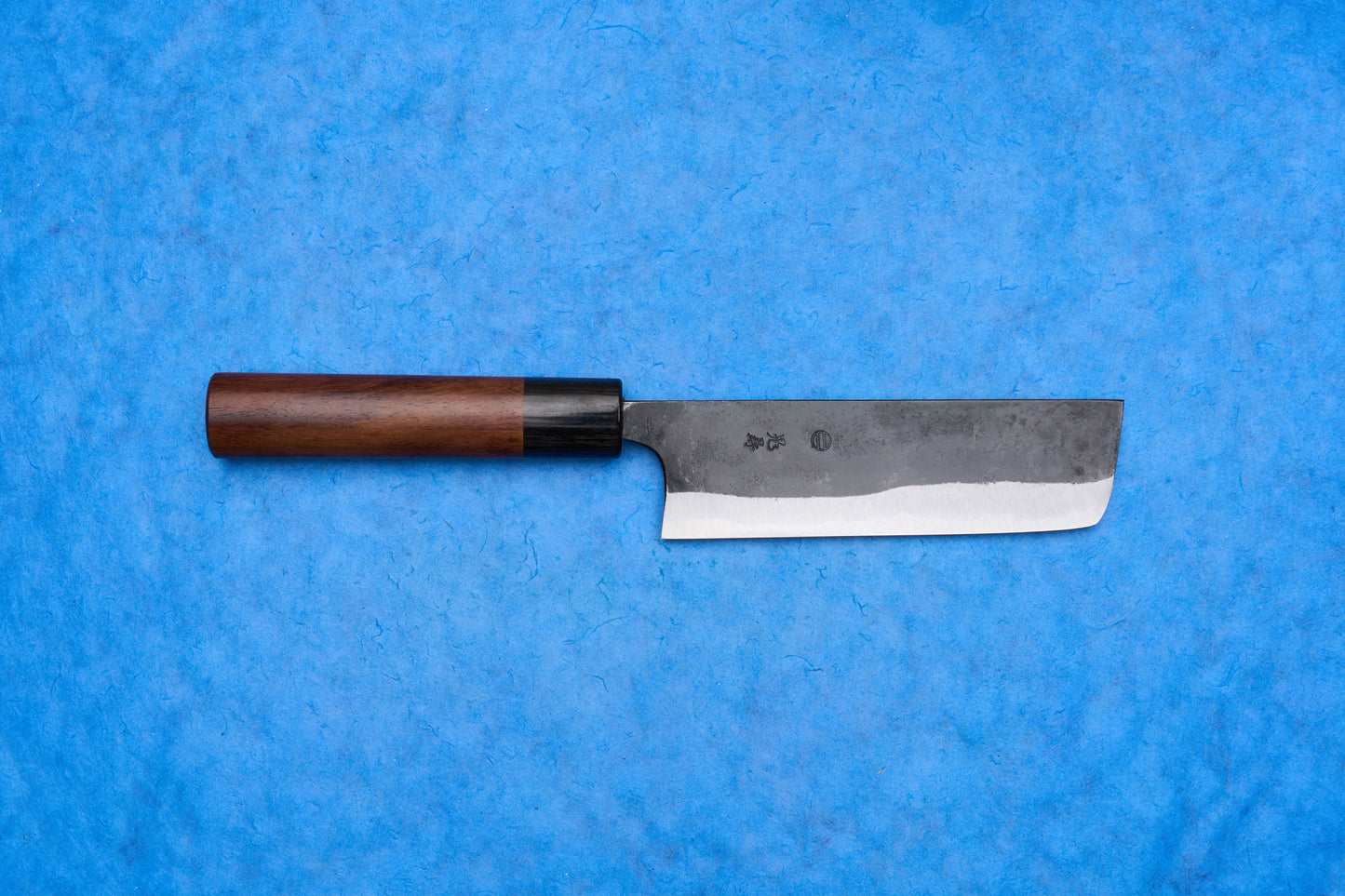 Onihiro Shiroichi Kurouchi Nakiri 135mm, a Japanese-style vegetable knife with a rectangular dual-tone blade and wooden handle, ideal for precise push/pull chopping, displayed on a blue background.