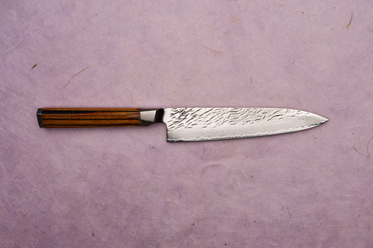 Ono Knife Suzukaze SG2 Tsuchime Damascus Petty 150mm showcases a hammered-finish Damascus blade with a striped wood handle, embodying precision craftsmanship on a pale lavender-pink textured background.