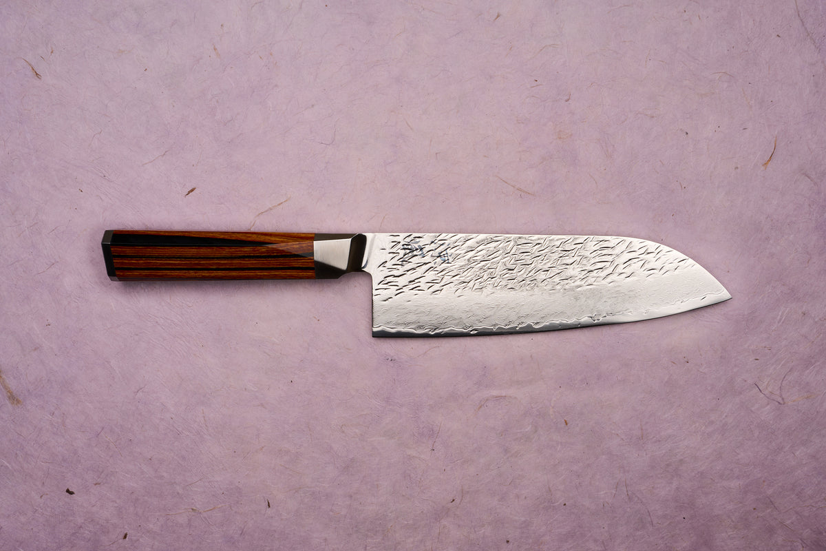 Ono Knife Suzukaze SG2 Tsuchime Damascus Santoku 180mm displayed on a textured surface, showcasing its hammered Damascus-style blade and dark octagonal wooden handle, embodying precision craftsmanship from Knifewear.