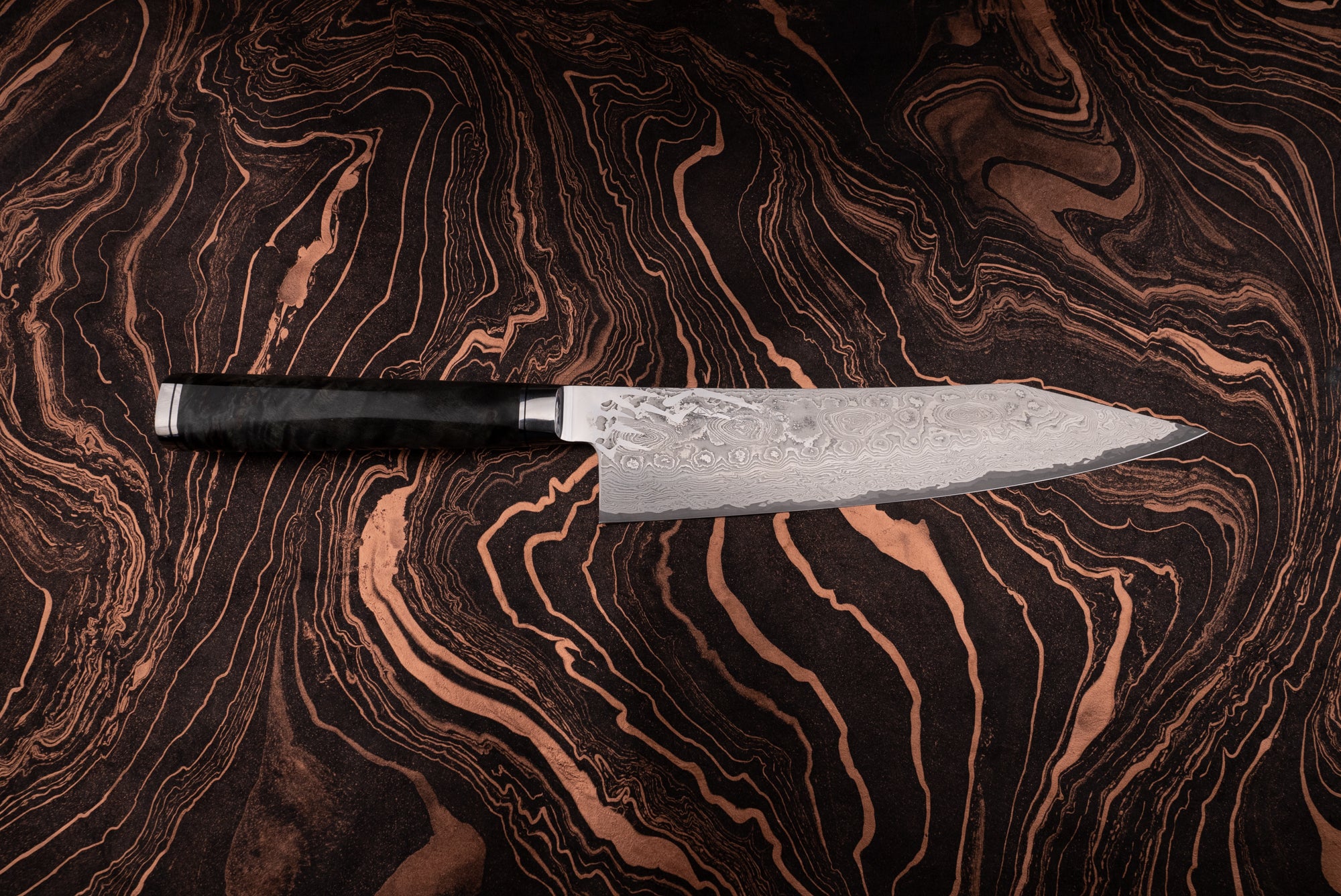 Ryusen Oukokuryu Gyuto 210mm knife with a Damascus-patterned blade and dark handle, showcased on a marbled surface echoing the knife's intricate design, embodying high-end Japanese craftsmanship.
