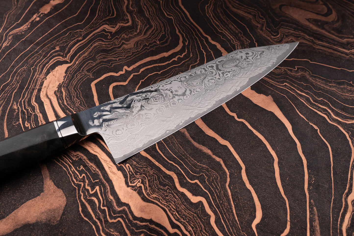 Close-up of a Ryusen Oukokuryu Gyuto 210mm with a Damascus-style blade featuring a swirling pattern, polished bolster, and dark handle, resting on a copper-accented dark surface.