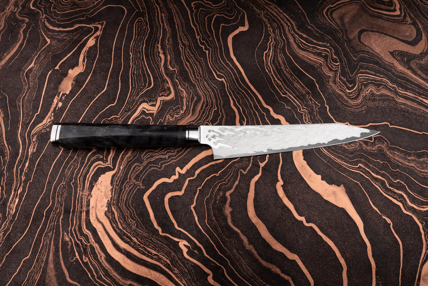 Ryusen Oukokuryu Petty 135mm knife with a Damascus-steel blade and a dark handle, showcased on a rich, patterned surface, epitomizing high-end Japanese craftsmanship and precision for smaller kitchen tasks.