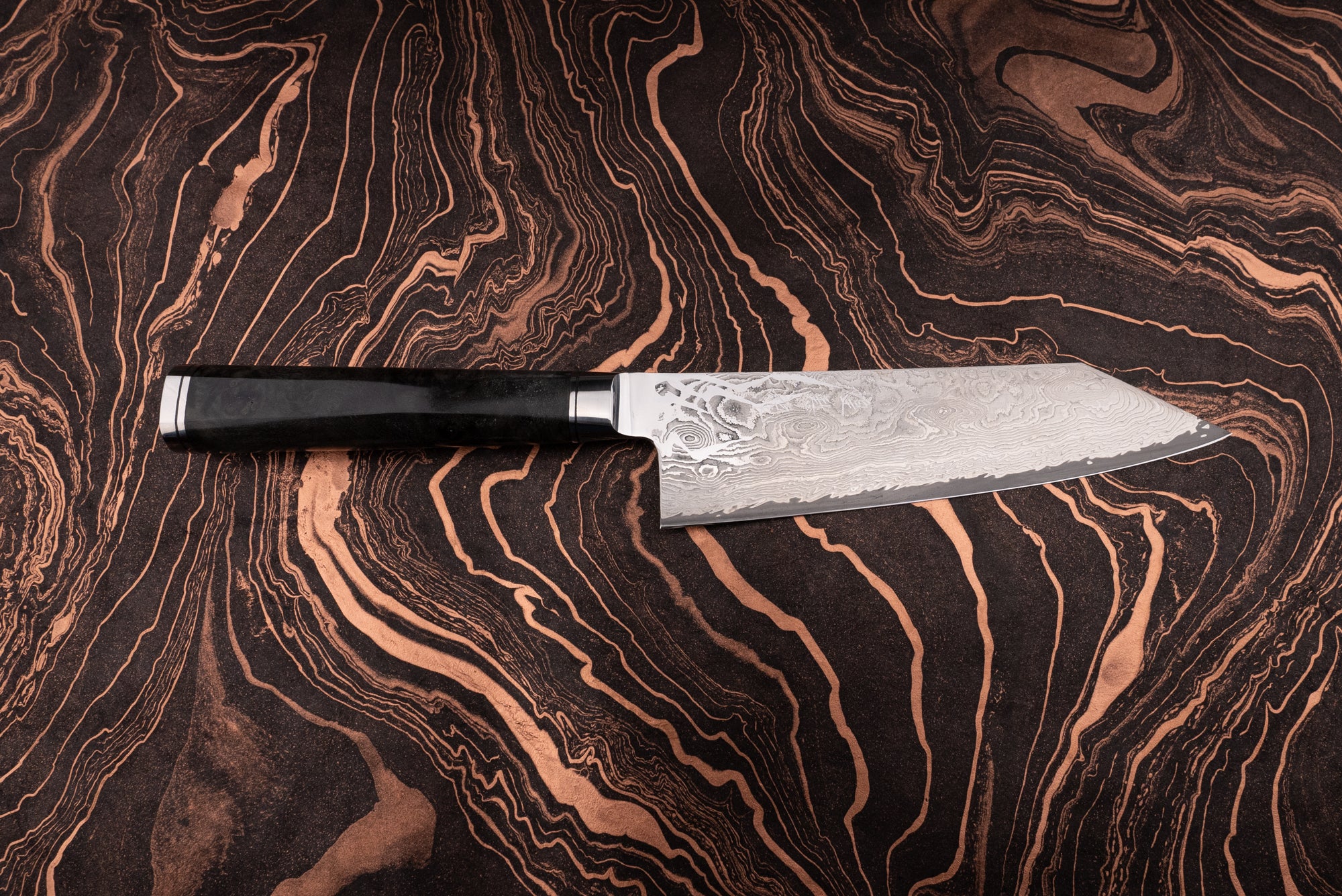 Ryusen Oukokuryu Santoku 170mm knife with a Damascus-style blade showcasing intricate layered patterns and a sleek black handle, resting on a dark, copper-streaked surface.