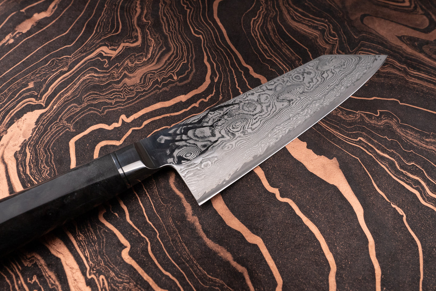 Ryusen Oukokuryu Santoku 170mm knife with a wavy Damascus-steel blade on a marbled surface; features a dark, ornate handle, epitomizing Japanese craftsmanship with superior edge retention and sleek design.