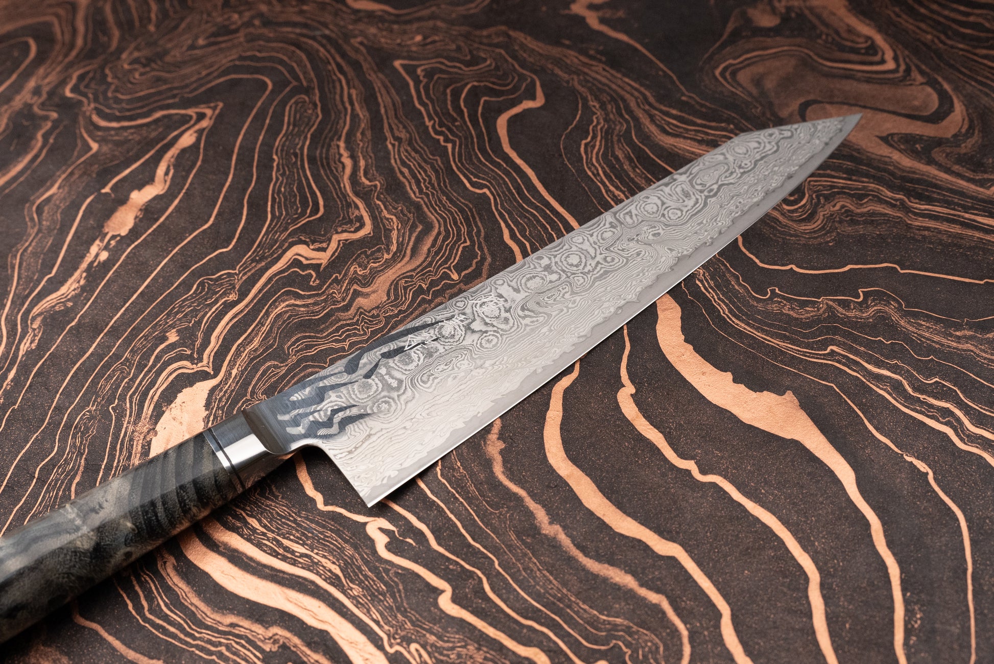 Ryusen Oukokuryu Sujihiki 260mm, a Damascus steel kitchen knife, features a swirling patterned blade and a resin-infused handle, ideal for precise slicing, showcased on a textured surface.