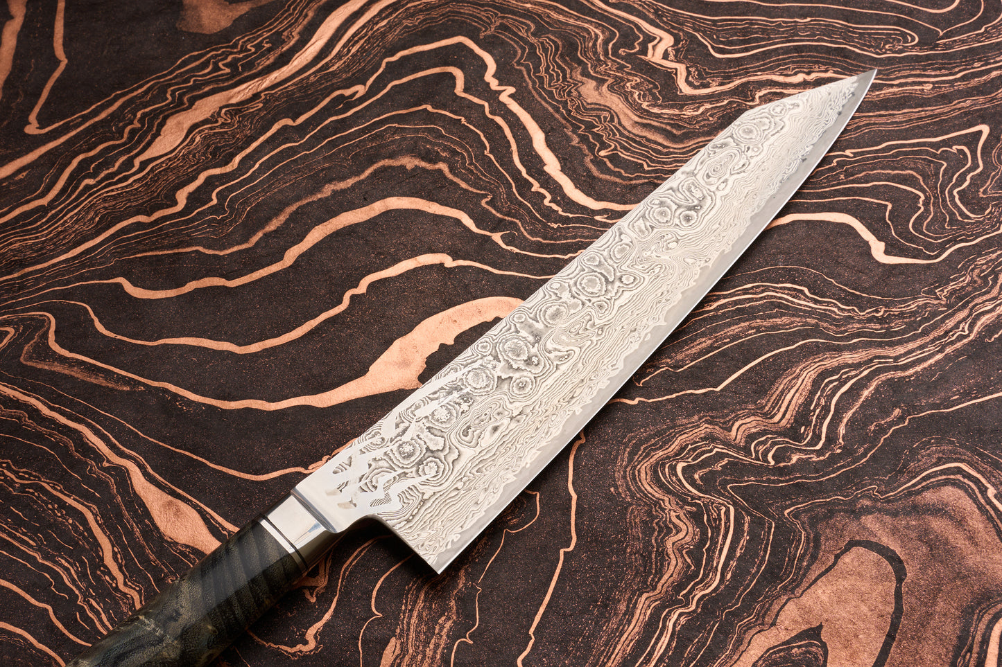 Ryusen Oukokuryu Gyuto 240mm knife with a Damascus-style steel blade rests on a marbled surface. The ornate handle's edge is visible, highlighting its craftsmanship and premium design.