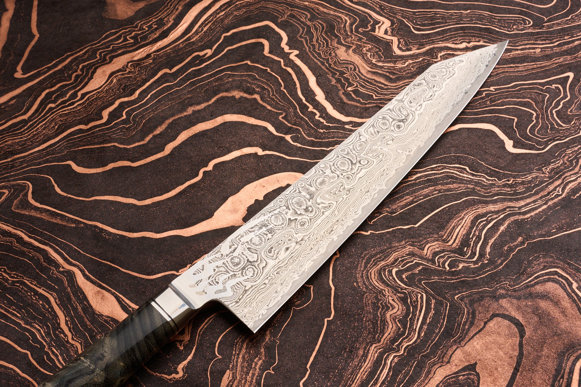 Ryusen Oukokuryu Gyuto 240mm knife with a Damascus-style steel blade rests on a marbled surface. The ornate handle's edge is visible, highlighting its craftsmanship and premium design.