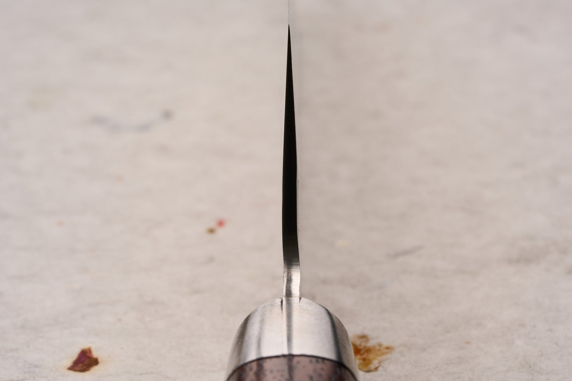 Sakai Takayuki SG2 Tsuchime Damascus Western Kengata Santoku 160mm knife stands upright, showcasing its narrow steel spine and metal bolster, with a glimpse of the wooden handle, on a cloth-dotted surface.