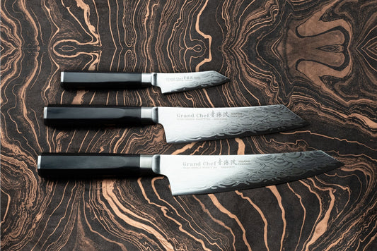 Sakai Takayuki Grand Chef Seigaiha 3 Piece Knife Set on wood-grain surface, featuring octagonal handles and damascus-style blades with Grand Chef stamp; sizes range from utility to chef’s knives.
